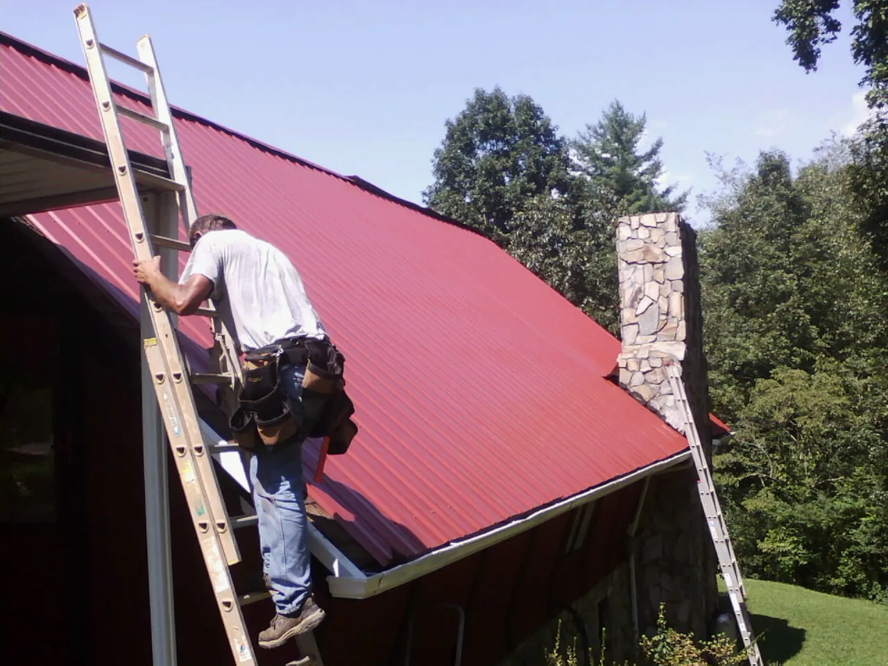 Roofer on a ladder beside a red metal roof and stone chimney — Soffit & Fascia Repair by Roofing Brothers in Baden